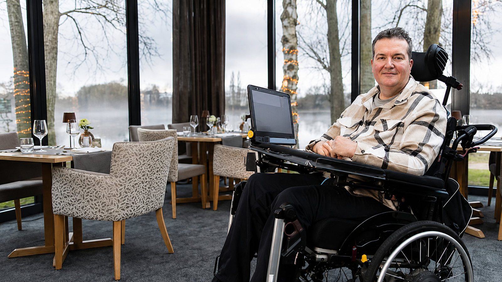 Andreas sitzt in einem elektrischen Rollstuhl mit seinem TD I-Serie in einem modernen Restaurant vor großen Fenstern mit Blick auf einen nebligen See.